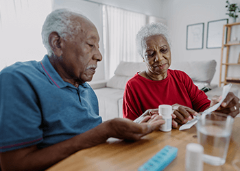 Gray-haired, Black couple reading instructions from a prescription bottle, with a pill organizer on the table.