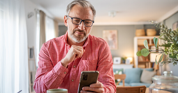 An older man looking at his phone while drinking a cup of coffee in his living room.