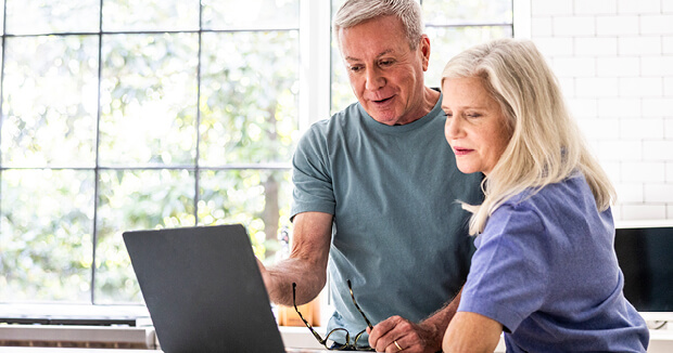 An older man and woman looking at computer on their desk with a sunny window behind them.