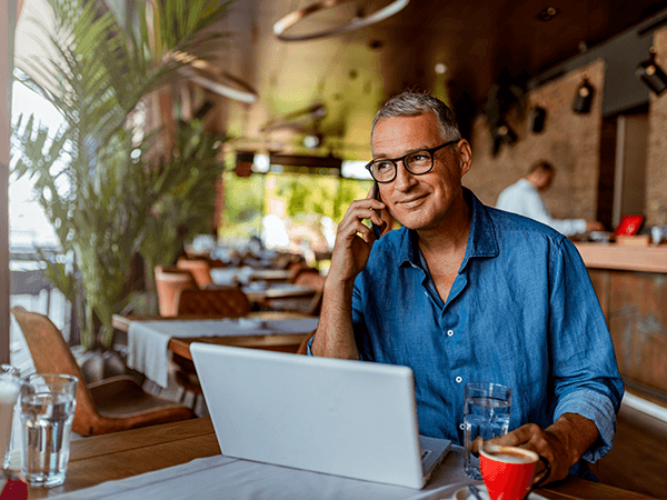 Businessman sitting in a tropical cafe, talking on his mobile phone, and working on a laptop.