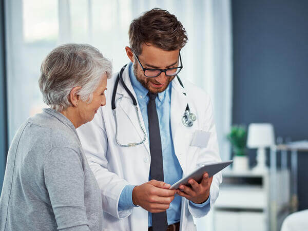 An elderly woman and a male doctor wearing a white coat and a stethoscope around his neck are both looking at an electronic tablet together