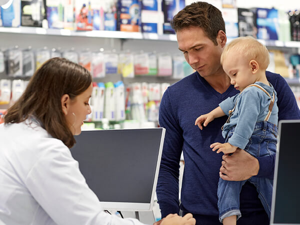 Female pharmacist explaining medicine to customer standing with baby boy. Male is carrying son while standing at checkout counter in store. They are in pharmacy.