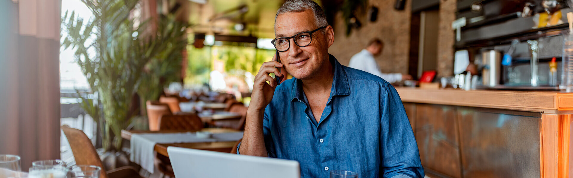 Gray-haired man with glasses smiling while using his phone and laptop in a coffee shop