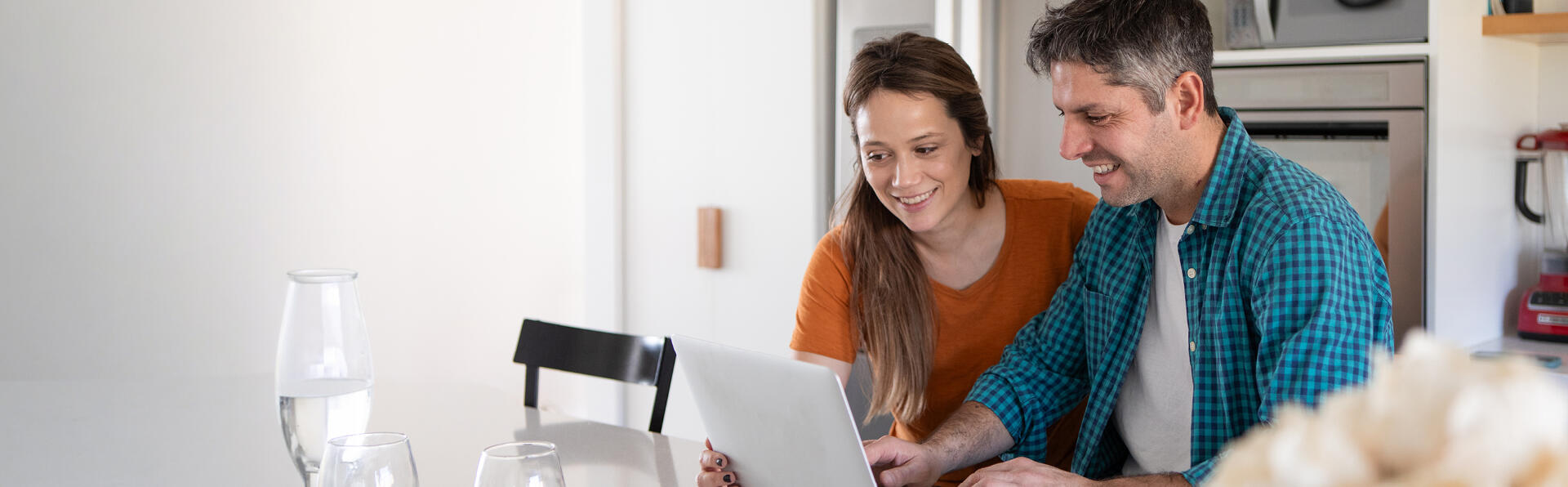A husband and wife in their kitchen looking at their computer screen and smiling.