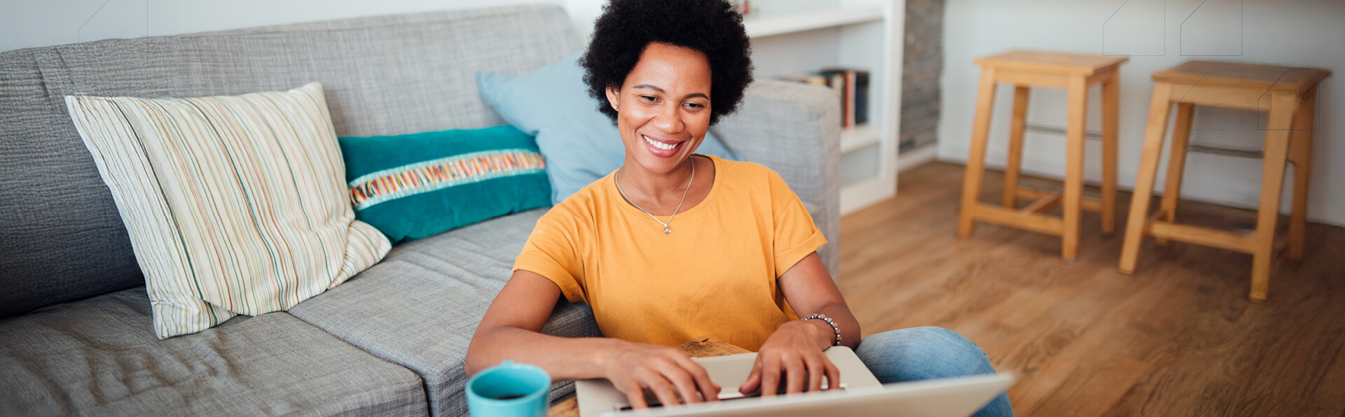 A young woman in her living room working on her laptop in front of her couch, smiling with a cup of coffee.