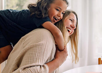 Child riding on an adult's back in a bright home dining area, with a laptop on the table nearby.