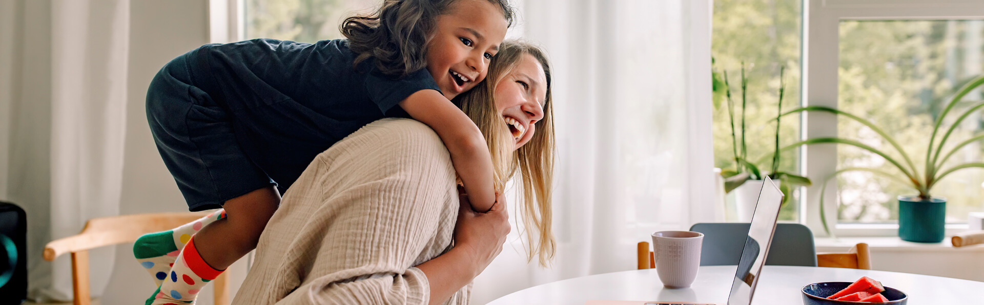 Child riding on an adult's back in a bright home dining area, with a laptop on the table nearby.