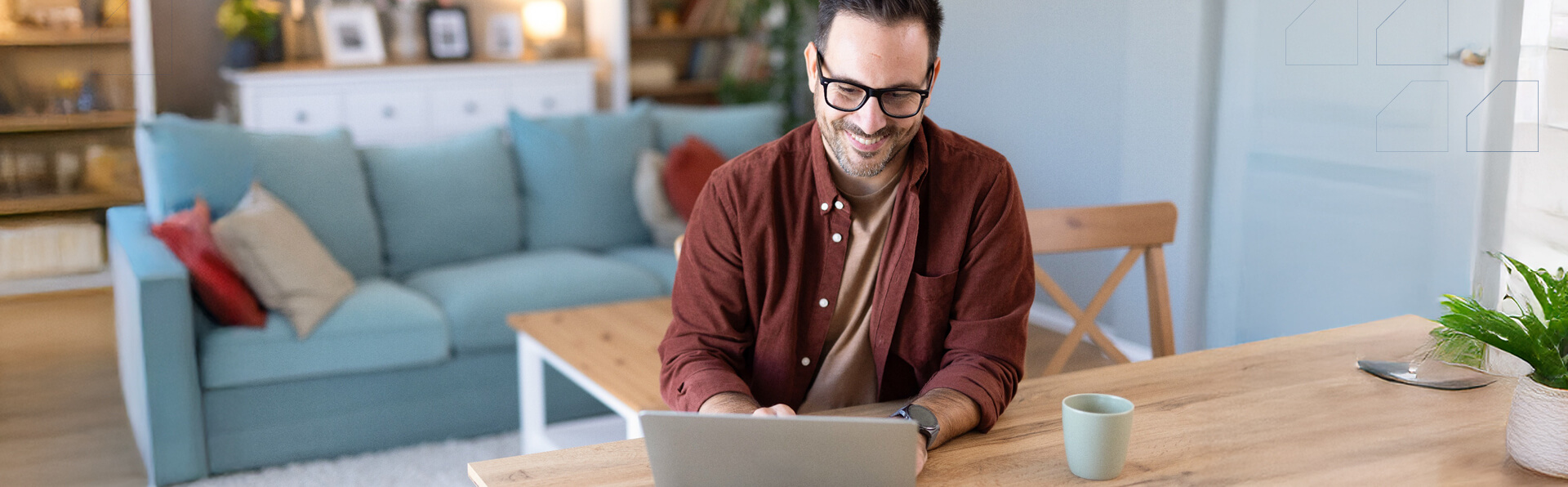 An approachable man with a cup smiles while working on his laptop at a tidy table, reflecting the ease of flexible work from home.