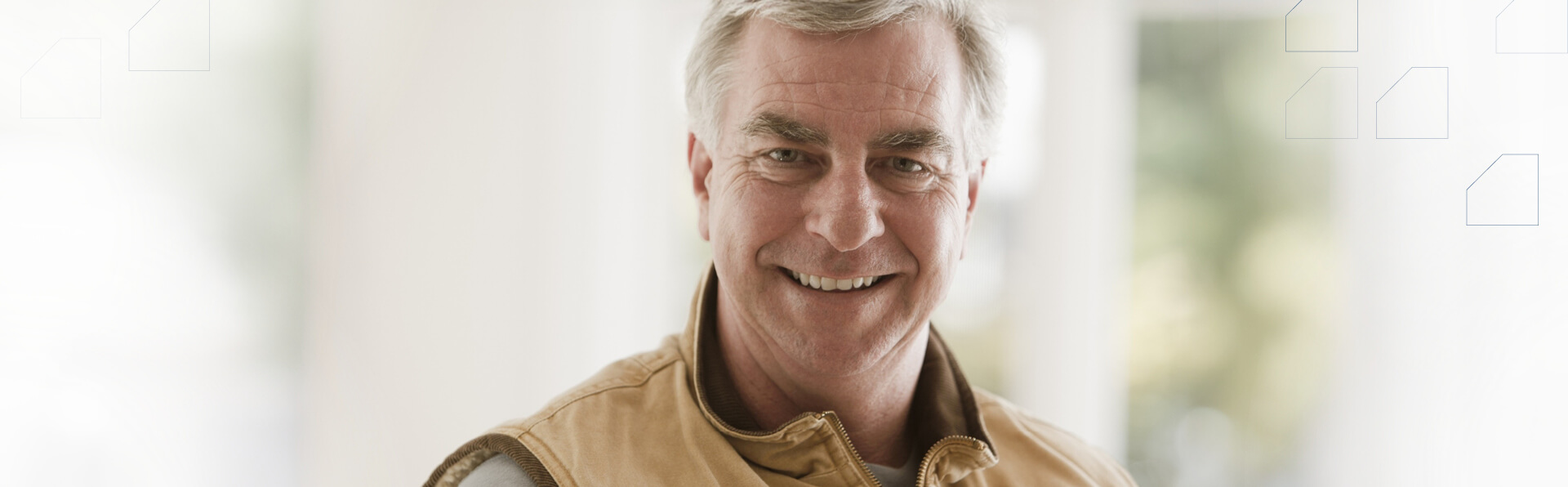Wearing a long-sleeved shirt and vest, a gray-haired Caucasian male smiles while a laptop sits on his lap