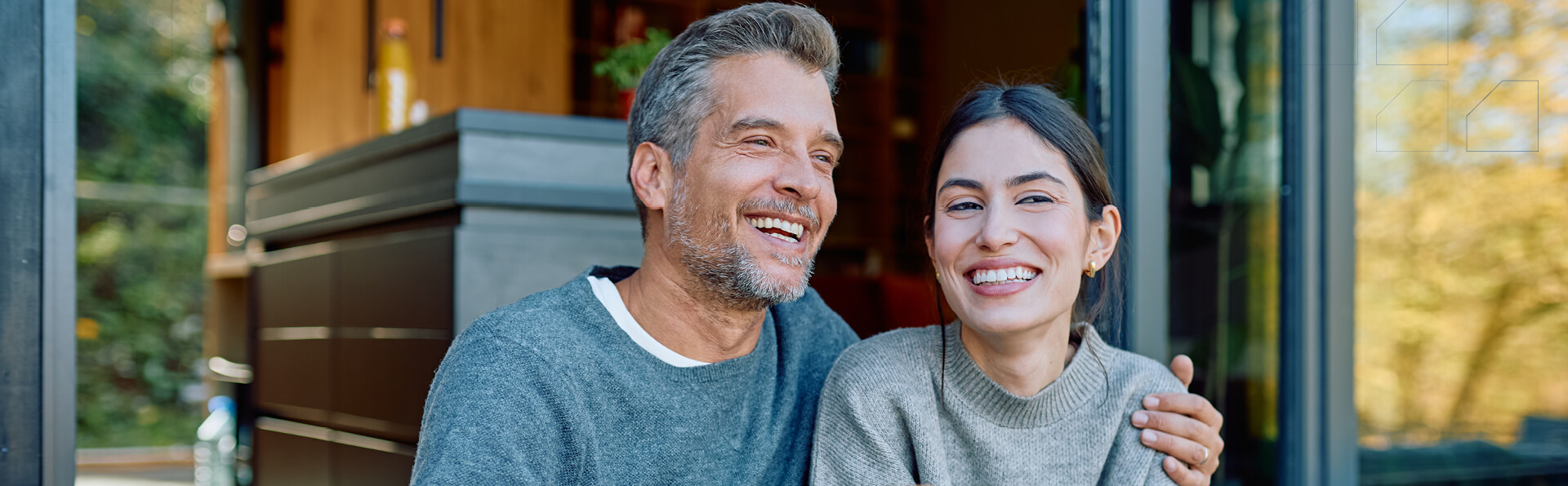 Cheerful couple relaxes, sitting on a wooden porch outside their house, sharing a joyful moment with hot drinks