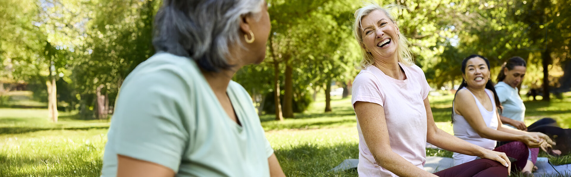 Happy woman sitting for yoga with friends. Mature female is laughing while enjoying morning routine. She is in public park.